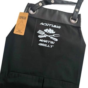 Op de afbeelding: Zwarte canvas schort met een leren bovenkant en verstelbare banden. De schort heeft een grote zak aan de voorkant en een kleinere zak aan de zijkant. De schort is gepersonaliseerd met de tekst "ACHTUNG MARTIN GRILLT" en een afbeelding van gekruiste grillgereedschap en een vlam.
