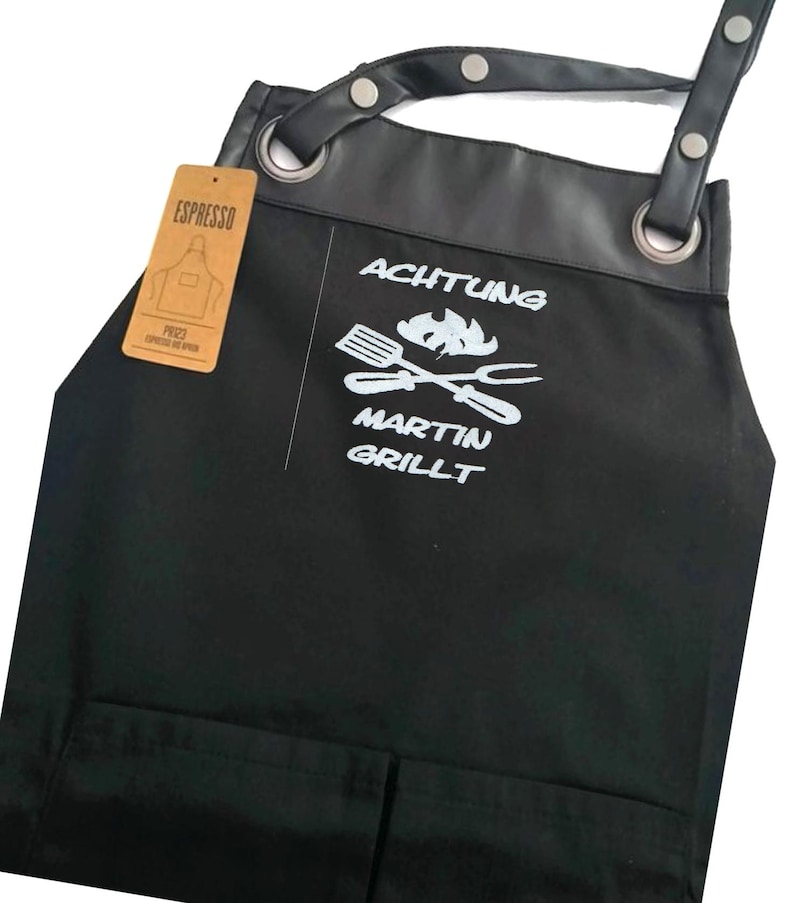 Op de afbeelding: Zwarte canvas schort met een leren bovenkant en verstelbare banden. De schort heeft een grote zak aan de voorkant en een kleinere zak aan de zijkant. De schort is gepersonaliseerd met de tekst "ACHTUNG MARTIN GRILLT" en een afbeelding van gekruiste grillgereedschap en een vlam.