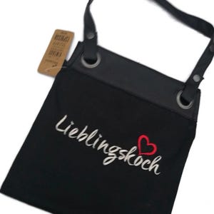 Op de afbeelding: Zwarte canvas schort met verstelbare banden en een grote zak. De schort heeft een wit geborduurd ontwerp met de tekst "Lieblingskoch" en een rood hart.