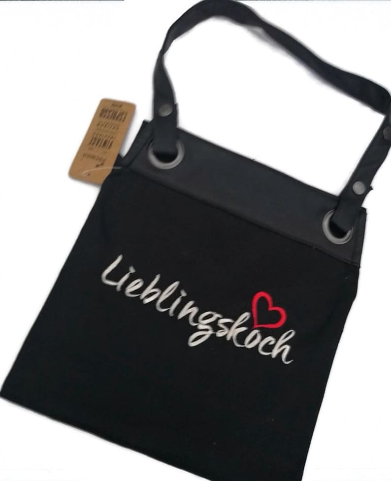 Op de afbeelding: Zwarte canvas schort met verstelbare banden en een grote zak. De schort heeft een wit geborduurd ontwerp met de tekst "Lieblingskoch" en een rood hart.