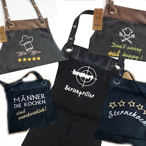 Op de afbeelding: Vijf verschillende zwarte schorten met verschillende geborduurde ontwerpen. De ontwerpen omvatten "Big Boss" met vijf sterren, "Don't worry eat happy!" met een koksmuts en gekruiste lepels, "M&auml;nner die kochen sind unwiderstehlich!" (Mannen die koken zijn onweerstaanbaar!), "bratort Seriengriller" (Grillmeester) en "Sternekoch" (Sterrenkok) met vijf sterren.