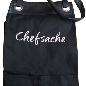 Op de afbeelding: Zwarte canvas schort met verstelbare banden en een grote zak. Het schort heeft het woord "Chefsache" in wit geborduurd op de voorkant.
