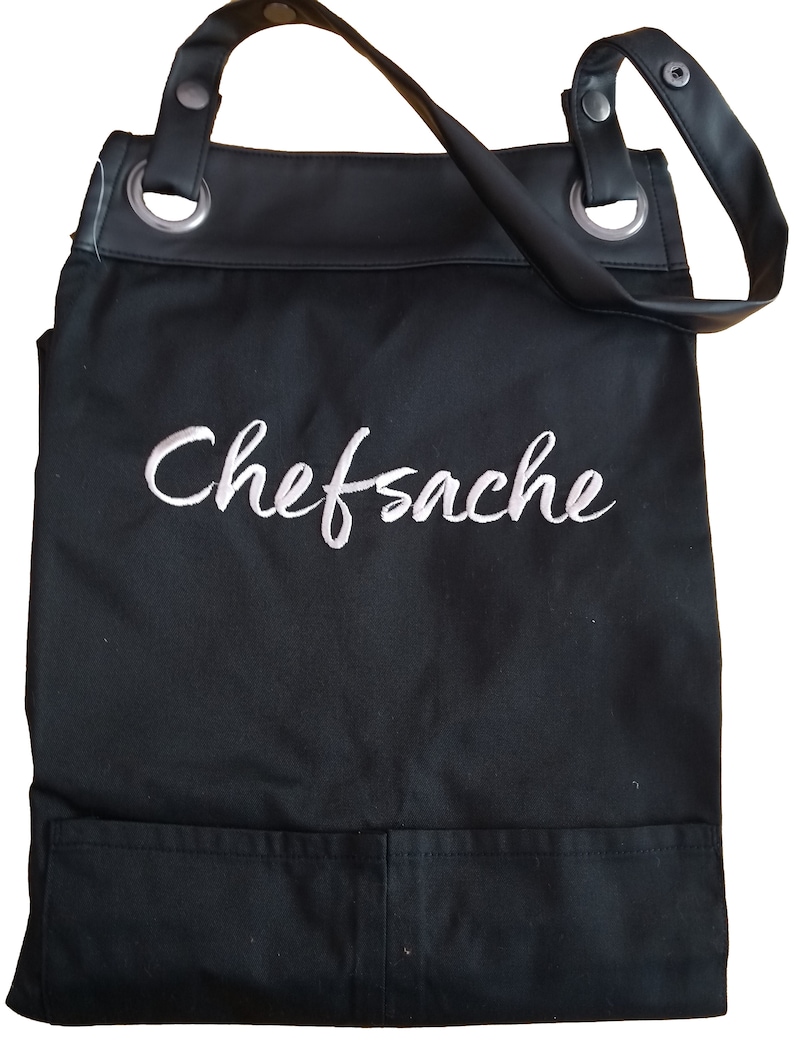 Op de afbeelding: Zwarte canvas schort met verstelbare banden en een grote zak. Het schort heeft het woord "Chefsache" in wit geborduurd op de voorkant.