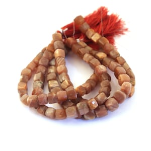 May include: Strands of faceted, cube-shaped beads in shades of orange and brown. The beads are strung together, with a red tassel at the top. The beads appear to be made of a stone material.