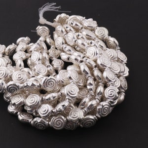 May include: A strand of silver-toned beads with a spiral design. The beads are oval-shaped and have a smooth, shiny finish.
