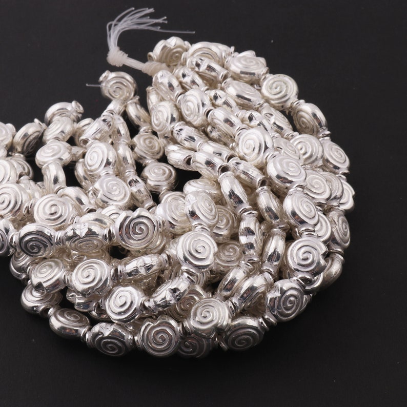 May include: A strand of silver-toned beads with a spiral design. The beads are oval-shaped and have a smooth, shiny finish.