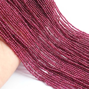 May include: Strands of small, round, deep red beads. The beads are tightly strung together, creating a flowing, textured appearance. The beads are a rich, dark red color, and appear to be made of a polished material. The image is well-lit, highlighting the beads' details.
