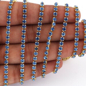 May include: A close-up of a gold chain with blue rhinestones. The chain is made of small, round rhinestones that are set in a gold metal setting. The chain is lying on a hand, and the rhinestones are sparkling in the light.