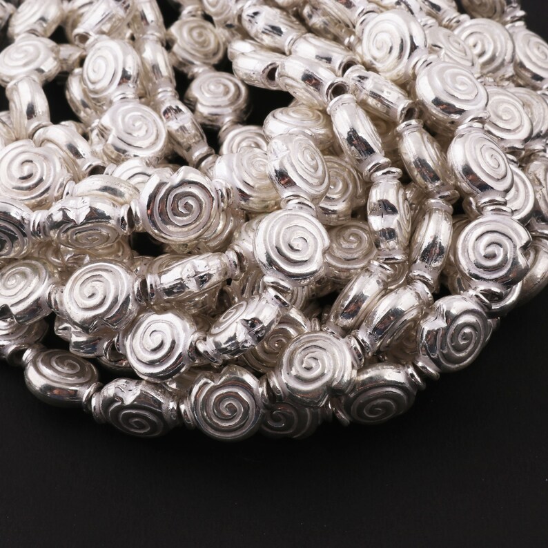 May include: A strand of silver-toned beads with a spiral design. The beads are arranged in a pile, showing the detail of the spiral pattern.
