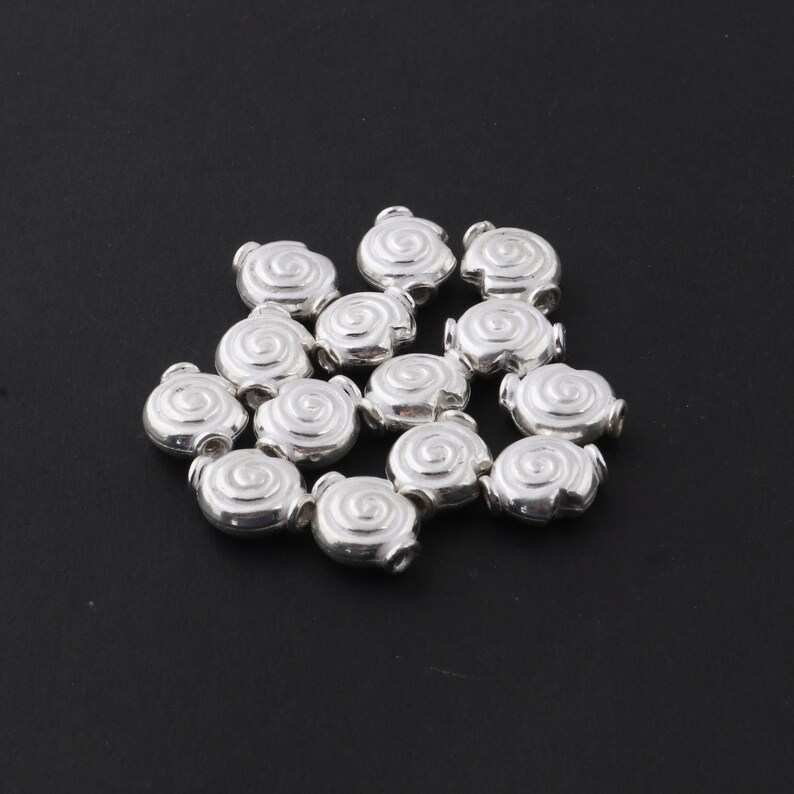 May include: A group of silver-toned metal beads with a spiral design. The beads are arranged in a loose pile on a black surface.