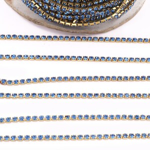 May include: A close-up of a gold chain with light blue rhinestones. The chain is laid out in a straight line on a white surface.
