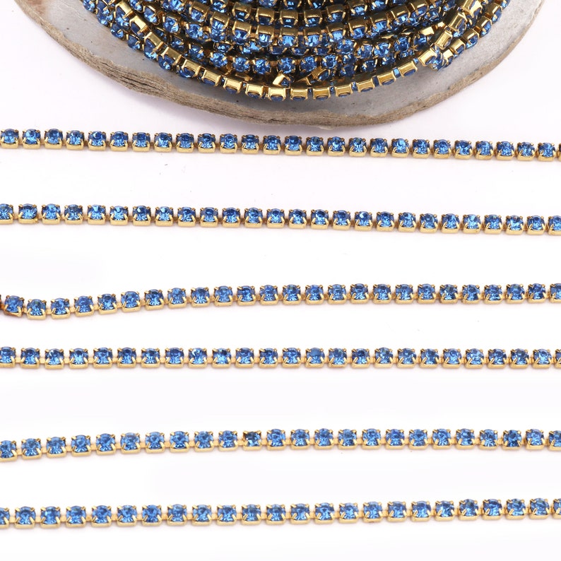 May include: A close-up of a gold chain with light blue rhinestones. The chain is laid out in a straight line on a white surface.