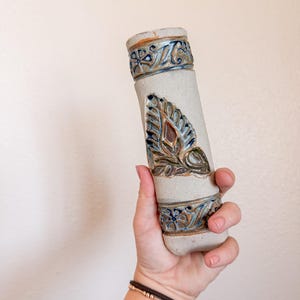 May include: A cylindrical ceramic vessel with a matte off-white finish, featuring two horizontal bands of embossed floral patterns and a central vertical leaf motif. The decorative elements are glazed in shades of blue, brown, and green.