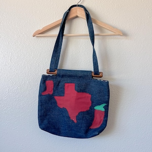 May include: A denim tote bag with a Texas theme. The bag features a red Texas outline, a red cowboy boot, and a red chili pepper with a green stem. The bag has blue straps and wooden handles.