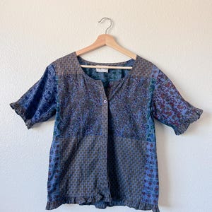 May include: A patchwork button-down shirt with short sleeves and a ruffled hem. The shirt features a mix of floral and geometric patterns in shades of blue, purple, and brown. The shirt is hanging on a wooden hanger.