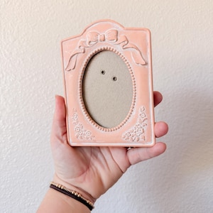 May include: A peach-colored picture frame with an oval opening. The frame has a bow at the top and floral details on the sides. The frame is held in a hand against a neutral background.