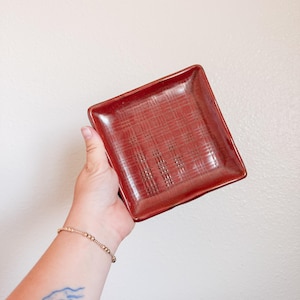 May include: A square ceramic dish with a glossy, deep red glaze. The surface features a subtle, impressed grid pattern of intersecting lines that create a textured, woven appearance across the center of the plate, finished with smooth, raised edges.