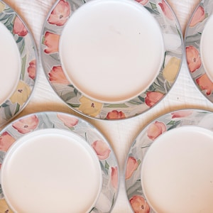 Peut inclure: Un ensemble de cinq assiettes rondes en céramique avec un centre blanc et une large bordure ornée d'un motif de tulipes roses et jaunes sur un fond de feuillage vert et gris atténué. Chaque assiette présente une finition lisse et brillante.