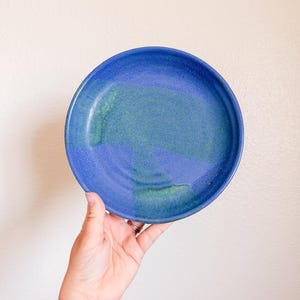 May include: A handmade ceramic plate with a vibrant blue glaze, featuring shades of teal and indigo. The plate is round and has a slightly raised edge. The plate is held by a hand.