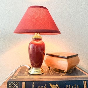 May include: A small table lamp with a red fabric shade and a glossy red base with a gold-tone metal bottom. The lamp sits on a dark blue book titled The Constitution of the United States of America, next to a small, light-colored wooden box.
