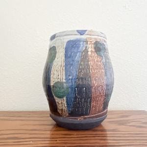 May include: A handcrafted ceramic vase with a textured surface. The vase features vertical stripes in blue, brown, and white, with green circular accents. The base is dark blue, and the top rim has a white and blue design. A unique piece for home decor.