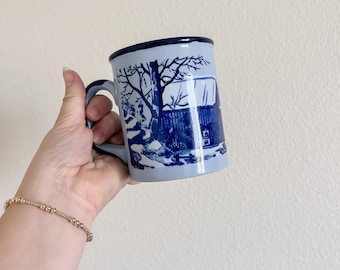 May include: A light blue ceramic mug with a dark blue rim and handle. The mug features a blue and white illustration of a winter scene with a tree and a building. The mug is held in a hand.