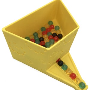 May include: Yellow plastic toy with a chute and a compartment filled with colorful translucent balls. The balls are red, green, yellow, and black.