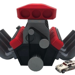 Puede incluir: Un modelo de motor V8 de gran tamaño impreso en 3D con detalles rojos y grises. El motor tiene ocho tubos de escape y una gran toma de aire. Un pequeño coche de juguete blanco está en primer plano.