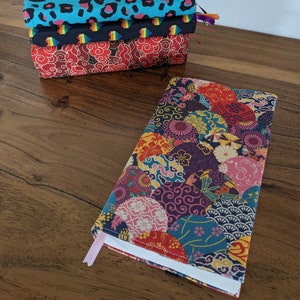 Può includere: Una pila di copertine per libri in tessuto con vari motivi. La copertina superiore è turchese con stampa leopardata, quella centrale ha un design arcobaleno e quella inferiore è rossa con un motivo floreale. Un libro con una copertina floreale colorata è aperto.