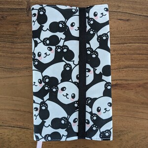Puede incluir: Una funda de libro de tela con estampado de panda blanco y negro con cierre el&aacute;stico negro. La funda tiene un marcap&aacute;ginas de cinta rosa.