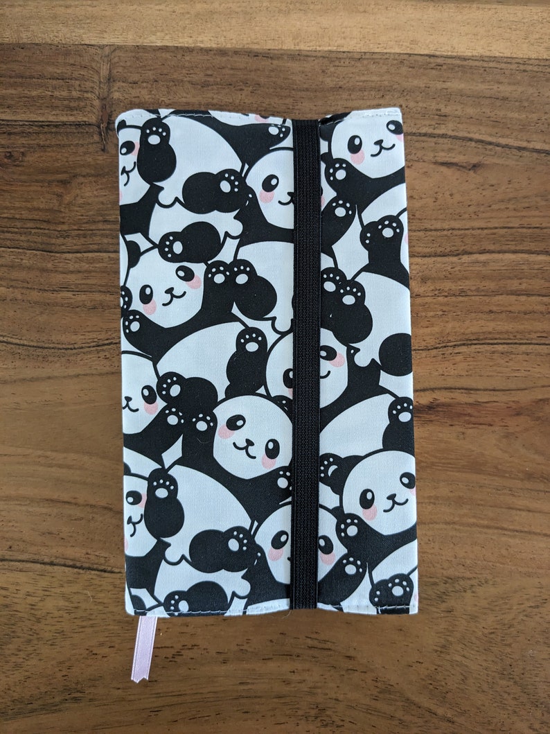 Puede incluir: Una funda de libro de tela con estampado de panda blanco y negro con cierre el&aacute;stico negro. La funda tiene un marcap&aacute;ginas de cinta rosa.