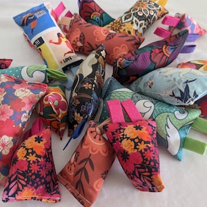 Puede incluir: Una colección de juguetes de tela para gatos llenos de hierba gatera, con varios patrones florales y geométricos. Los juguetes están llenos de hierba gatera y tienen un lazo para colgar.