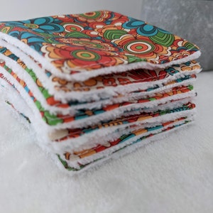 Puede incluir: Una pila de pa&ntilde;os cuadrados de tela con un estampado floral retro en tonos naranja, azul, verde y rojo. Cada pa&ntilde;o tiene un borde suave, blanco y esponjoso. Los pa&ntilde;os est&aacute;n apilados, mostrando la tela estampada y el borde blanco.