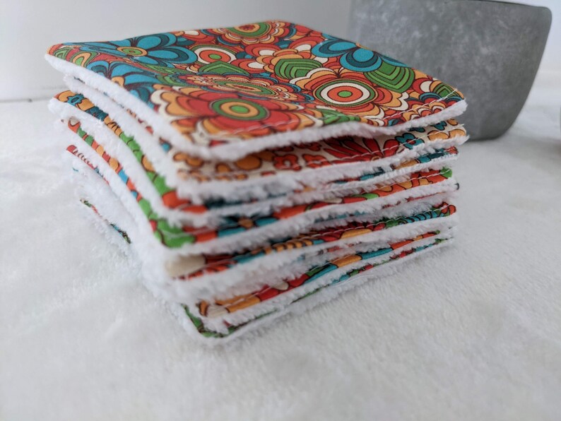 Puede incluir: Una pila de pa&ntilde;os cuadrados de tela con un estampado floral retro en tonos naranja, azul, verde y rojo. Cada pa&ntilde;o tiene un borde suave, blanco y esponjoso. Los pa&ntilde;os est&aacute;n apilados, mostrando la tela estampada y el borde blanco.