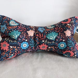 Puede incluir: Una almohada azul marino en forma de hueso con un estampado floral. El estampado incluye flores y hojas turquesas, coral y blancas. La almohada está diseñada para el soporte del cuello y se coloca sobre una superficie blanca.