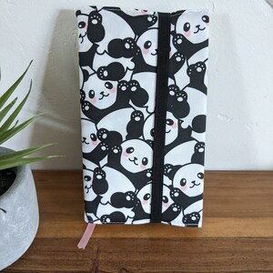 Puede incluir: Una cubierta de libro con estampado de panda blanco y negro con cierre el&aacute;stico negro y un marcap&aacute;ginas de cinta rosa.