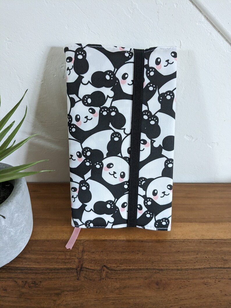 Puede incluir: Una cubierta de libro con estampado de panda blanco y negro con cierre el&aacute;stico negro y un marcap&aacute;ginas de cinta rosa.