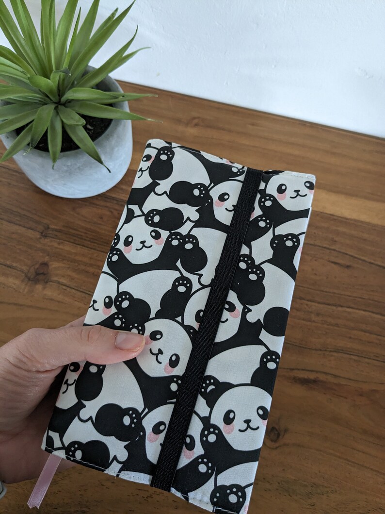 Puede incluir: Una funda para cuaderno con estampado de panda en blanco y negro con cierre el&aacute;stico negro. La funda est&aacute; hecha de tela y tiene un marcap&aacute;ginas de cinta rosa.