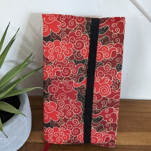 Puede incluir: Portada de cuaderno roja y dorada con un patrón de nubes en espiral. La portada tiene un cierre de banda elástica negra.