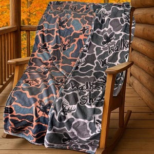 May include: Two camouflage-patterned blankets draped over a wooden rocking chair. One blanket features a gray, blue, and peach design with deer and bird silhouettes. The other blanket has a gray and white pattern with the text "Walk" and "Shack".