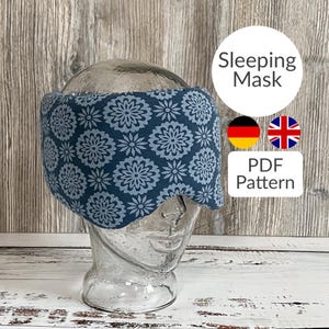 Könnte beinhalten: Eine blaue Schlafmaske mit weißem Blumenmuster. Die Maske befindet sich auf einer Glaskopfform. Der Text "Sleeping Mask" und "PDF Pattern" befindet sich auf dem Bild. Die Flaggen von Deutschland und Großbritannien sind ebenfalls auf dem Bild.
