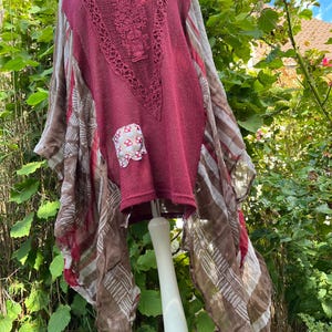 Könnte beinhalten: Eine weinrote Tunika mit Häkel-Ausschnitt und einem Pilz-Patch. Die Tunika hat lange, fließende Ärmel mit einem braun-weißen Streifenmuster. Das Kleidungsstück wird auf einer weißen Schaufensterpuppe in einem Garten gezeigt.