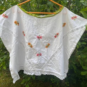 Könnte beinhalten: Eine weiße Kurzarmbluse mit weitem Ausschnitt und lindgrünem Saum. Das Kleidungsstück ist mit kleinen gestickten orangefarbenen und rosafarbenen Blumenmotiven verziert und verfügt über weiße Spitzeneinsätze sowie einen gewellten Spitzenabschluss.
