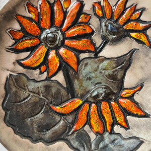 Könnte beinhalten: Ein Keramikteller mit einem stilisierten Design aus drei Sonnenblumen mit orangefarbenen und gelben Blütenblättern auf einem dunkelgrauen Hintergrund.