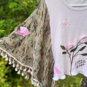 Könnte beinhalten: Ein hellrosa T-Shirt mit einem floral gemusterten Ärmel und Quastenbesatz. Die Vorderseite zeigt gestickte Blumen, rosafarbene Vichy-Stoffflicken und den Schriftzug "Love and Hope" in dunkelbrauner Schreibschrift auf der unteren rechten Seite.