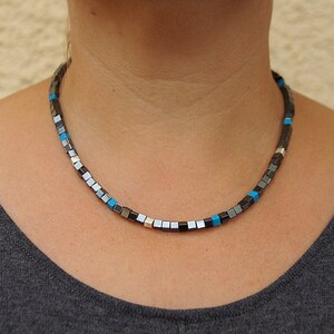Può includere: Una collana composta da piccole perline quadrate nei toni del grigio, argento e blu turchese. La collana, realizzata in materiale metallico, è indossata attorno al collo. Le perline sono uniformemente distanziate, creando un motivo geometrico.