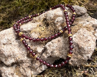Op de afbeelding: Een kralenketting met kleine, gefacetteerde, dieprode granaatkralen in een doorlopende streng. De ketting is geaccentueerd met gelijkmatig verdeelde, kleine, cilindervormige, goudkleurige metalen tussenstukjes voor een contrast.