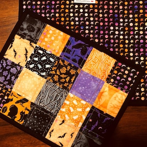 Puede incluir: Un juego de dos manteles individuales acolchados con temática de Halloween. El mantel individual más pequeño está hecho de tela de patchwork con varios motivos de Halloween, incluyendo murciélagos, fantasmas, calabazas y cuervos. El mantel individual más grande tiene un fondo negro con un patrón repetido de lunas y estrellas naranjas y rosas.