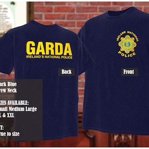 Może przedstawiać: Koszulka z ciemnoniebieskiego materiału z okrągłym dekoltem, na której z tyłu nadrukowano żółtym kolorem słowa "GARDA IRELAND'S NATIONAL POLICE". Z przodu koszulki znajduje się wyszywana odznaka w kolorze żółtym i niebieskim z napisem "IRELAND NATIONAL POLICE".
