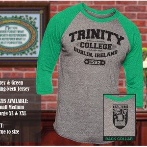 Könnte beinhalten: Ein grau-grünes Baseballtrikot mit einem weißen Ringkragen. Auf der Vorderseite des Trikots steht in schwarzer Schrift "Trinity College Dublin, Irland 1592". Der hintere Kragen ist mit einem schwarzen Aufdruck eines Wappens versehen, über dem das Wort "Trinity" steht.